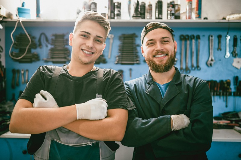 Werkshalle: Zwei jüngere Männer in Arbeitskleidung lächeln mit überkreuzten Armen in die Kamera