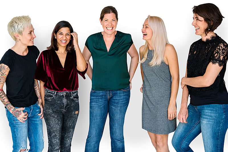 Fünf Frauen in Jeans und sommerlicher Kleidung stehen nebeneinander vor einer weißen Wand und schauen fröhlich in die Kamera